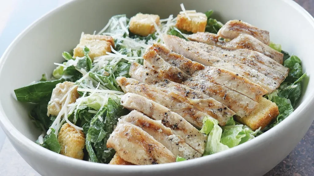 Caesar salad with grilled chicken, Parmesan, and croutons in white bowl.