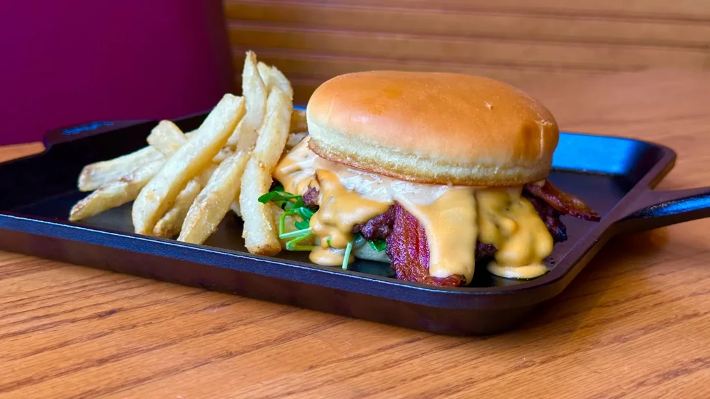 Burger with melted cheese, bacon, arugula, and golden fries on a skillet.