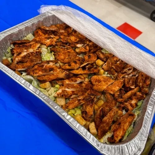 Foil tray of Caesar salad with grilled chicken, partially covered on blue.