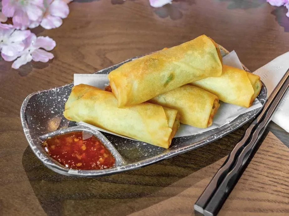 Golden-brown fried spring rolls, red chili sauce, and chopsticks on a table.