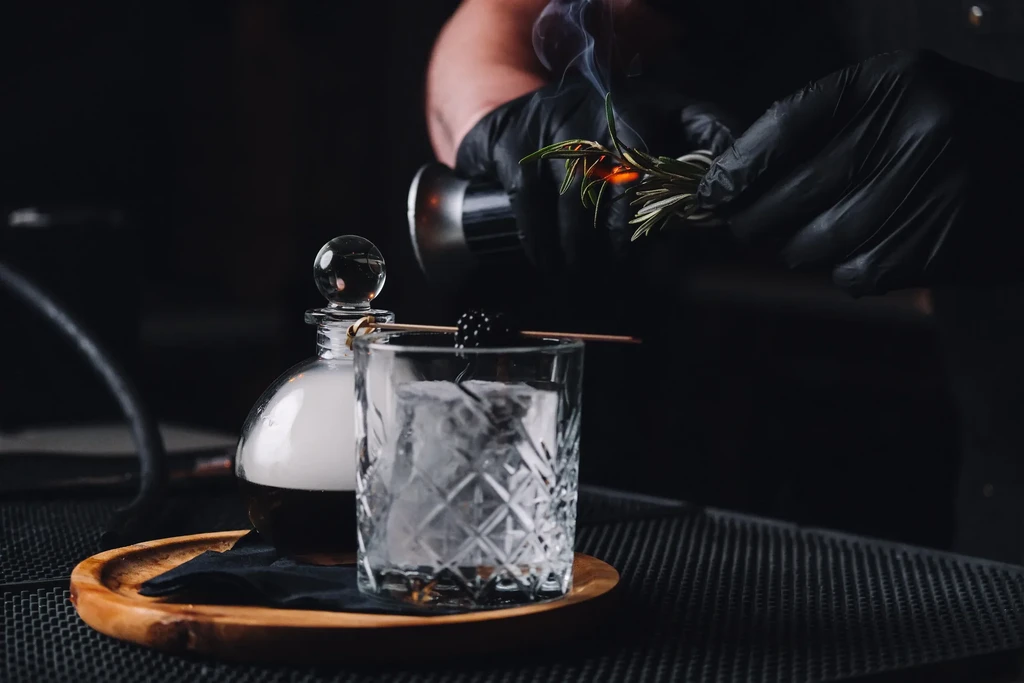 Gloved hand torches rosemary over a smoked cocktail with blackberry.