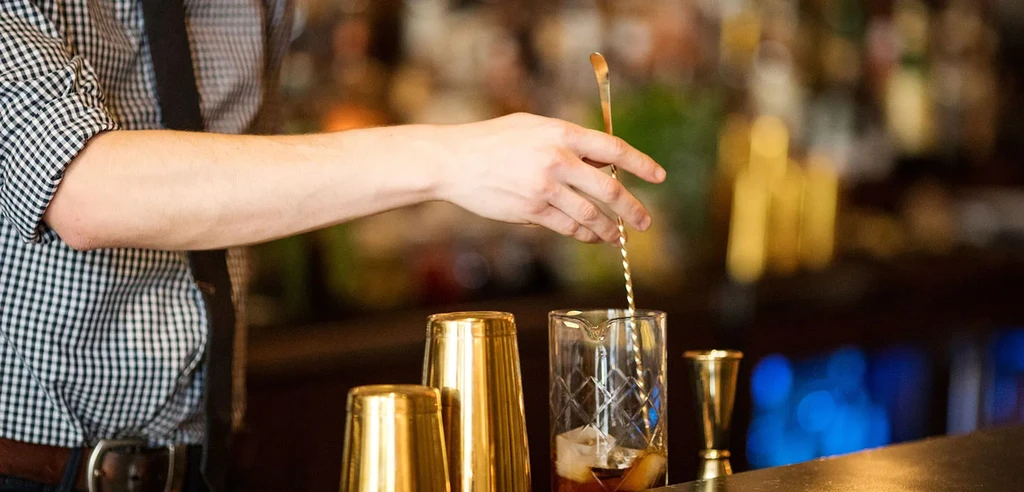 Bartender stirs a dark cocktail with a bar spoon in a mixing glass.