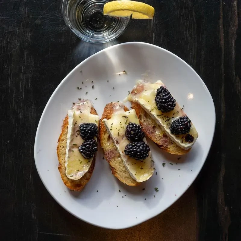 Plate of crostini with melted brie, blackberries, and herbs.