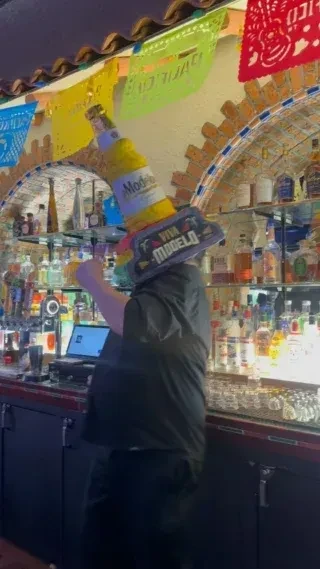 Bartender wearing a Modelo bottle-sombrero hat at a festive bar.
