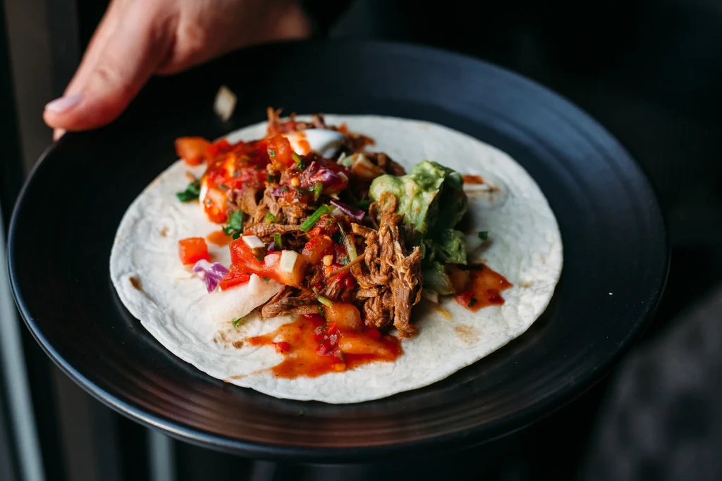 Hand holding a single meat taco with salsa, guacamole, and herbs.