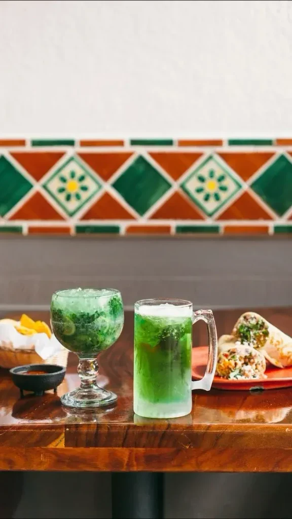Table with two green drinks, tortilla chips, red sauce, and burritos.