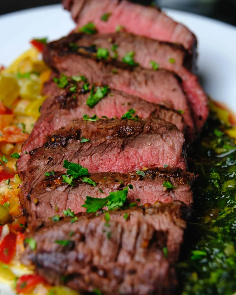 Plated medium-rare sliced steak with vegetables, herbs, and green sauce.