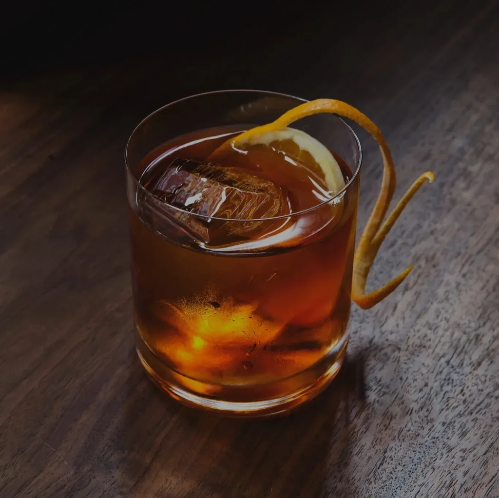 Amber cocktail with square ice and citrus peel on dark wood.