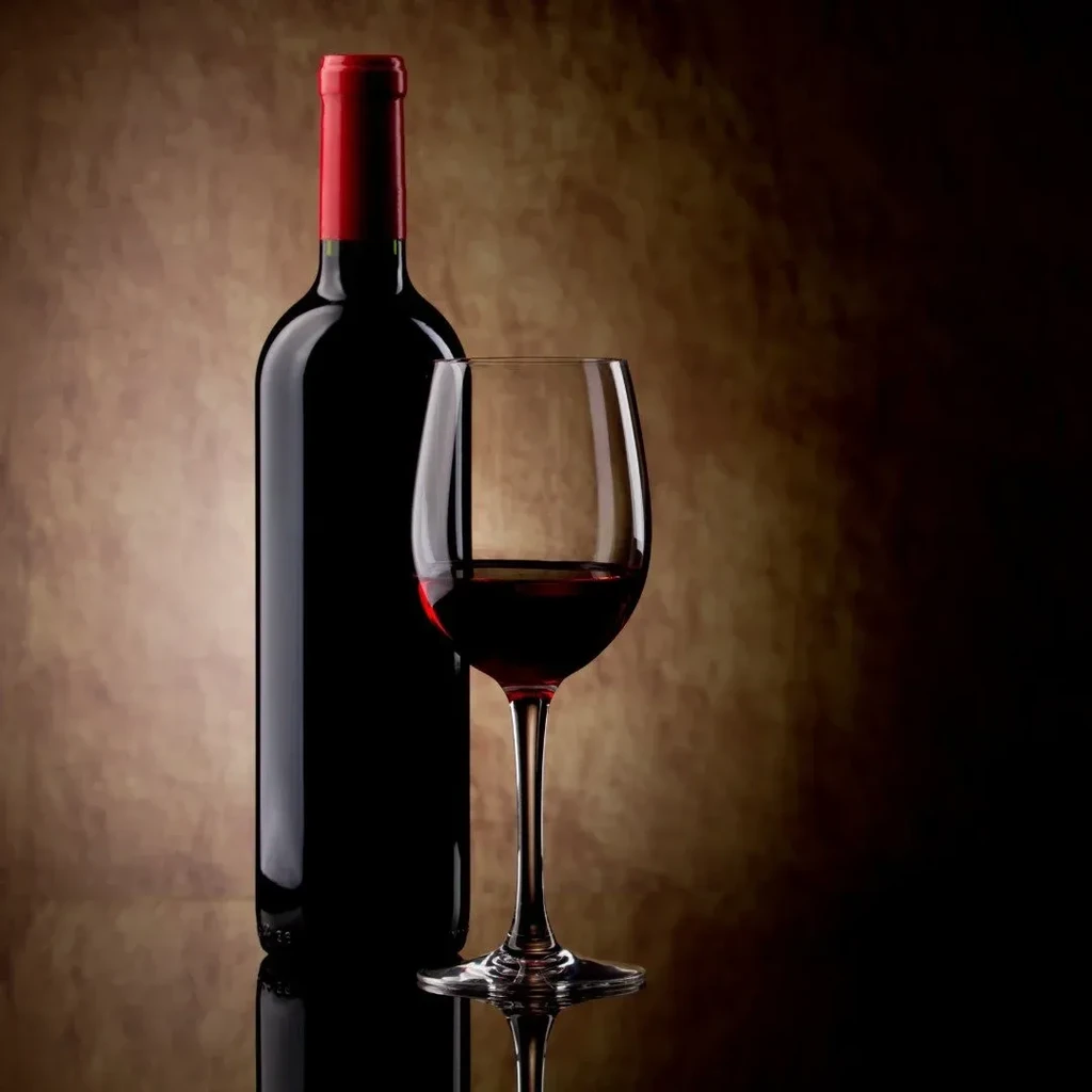 Red wine bottle with red foil, full wine glass on reflective surface.
