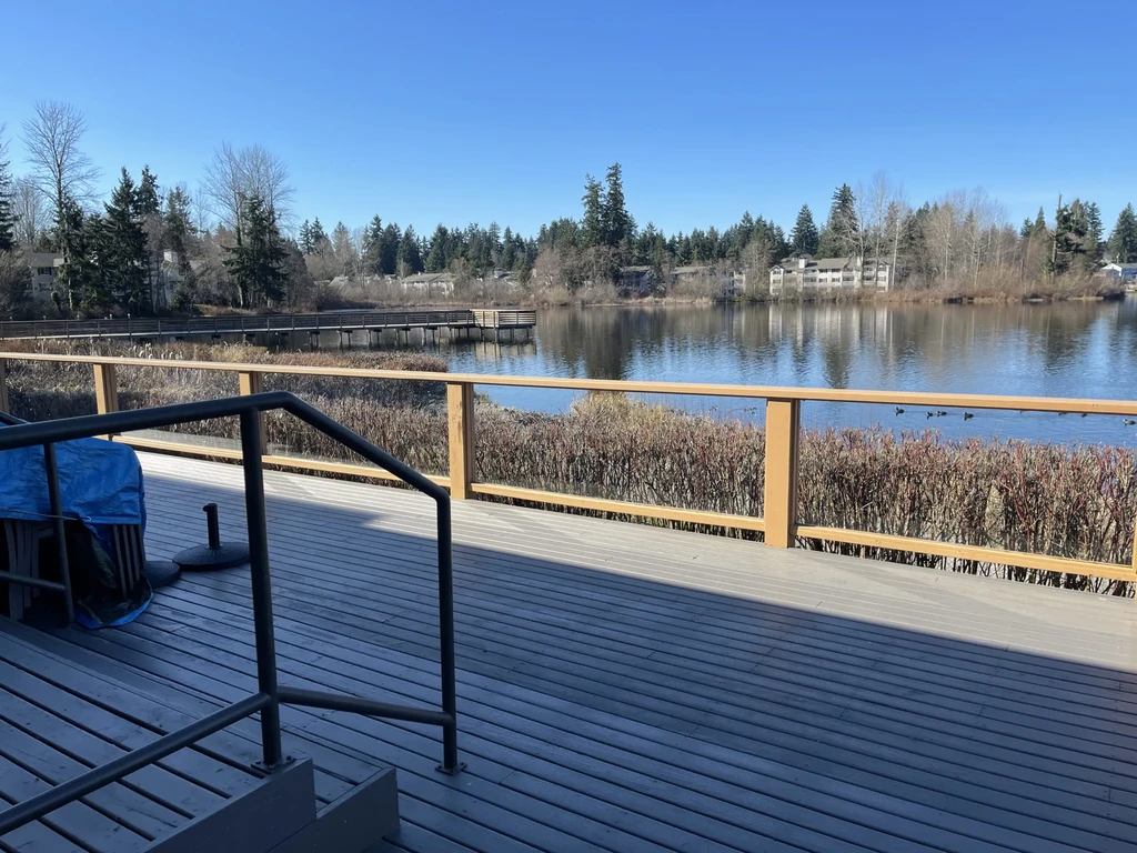 Wooden deck with glass railing overlooks a calm lake on a sunny day.