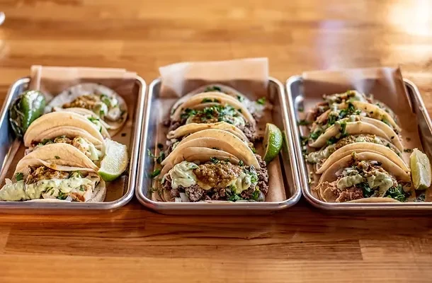 Three metal trays of tacos with various toppings on a wooden table.