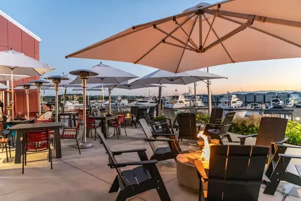 Restaurant patio with fire pits and tables, overlooking a marina at dusk.