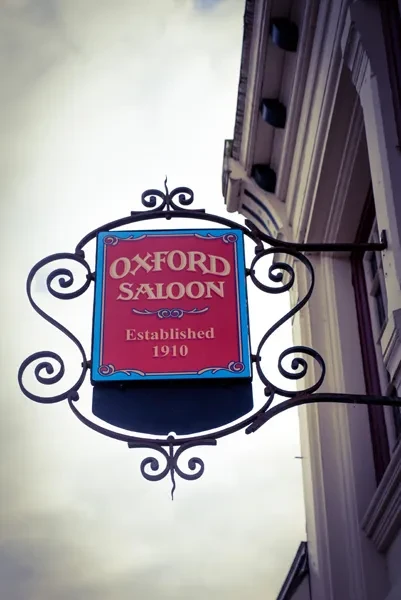 Ornate vintage sign for Oxford Saloon, established 1910, against cloudy sky.