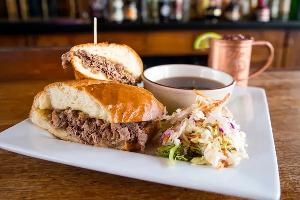 Beef dip sandwich, coleslaw, and au jus on a plate at a bar.