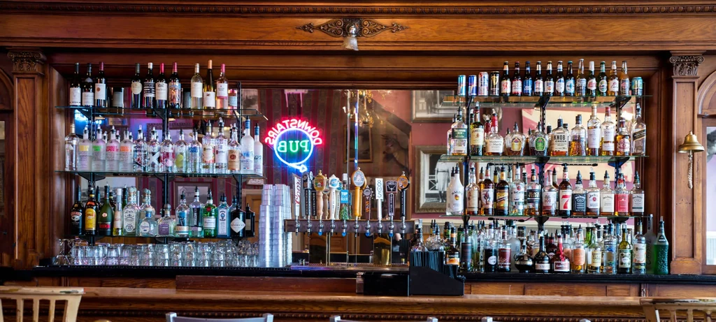 A fully stocked wooden bar reflects a 'DOWNSTAIRS PUB' neon sign.