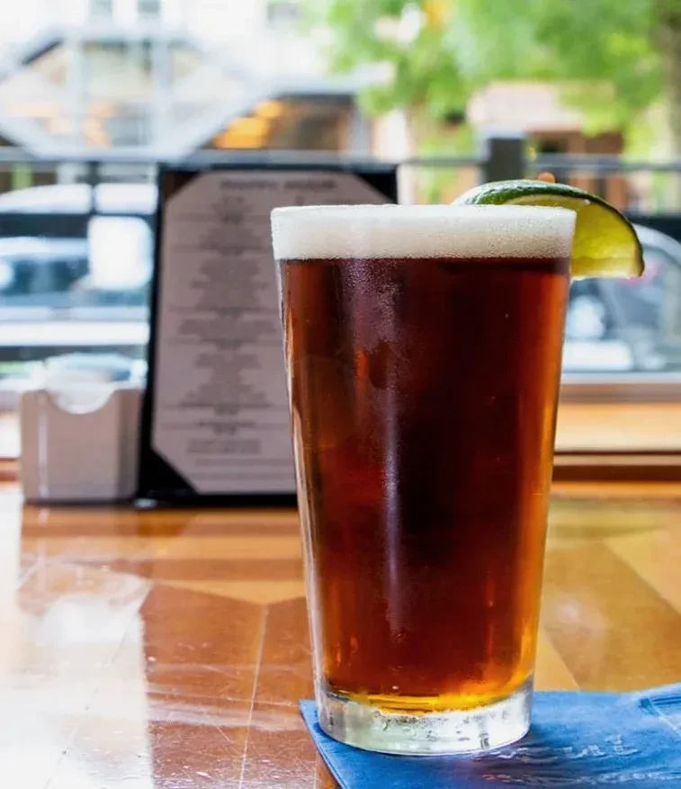 Cold reddish-brown beverage with frothy foam and lime on wood.