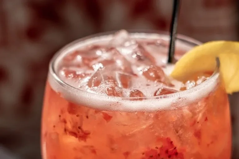 Coral drink with ice, muddled fruit, foam, straw, and lemon.