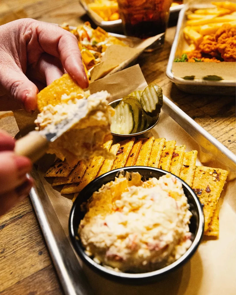 Close-up of hand scooping dip onto cracker next to pickles.