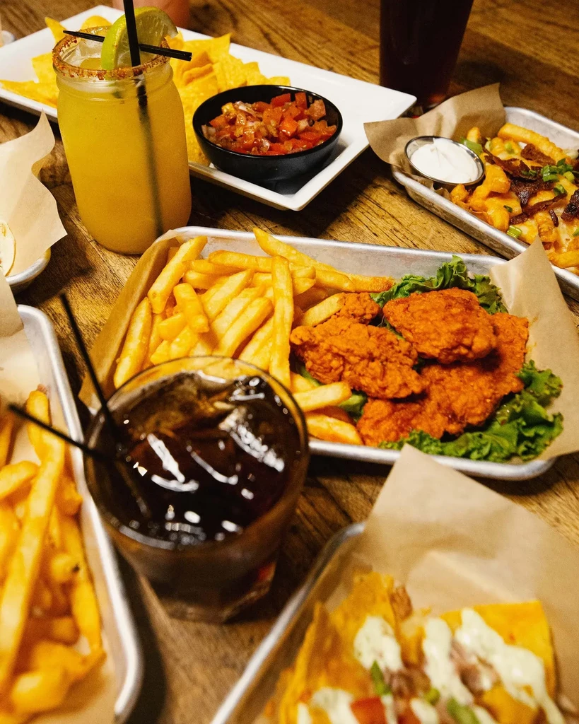 Table with fried chicken, loaded fries, nachos, salsa, and beverages.