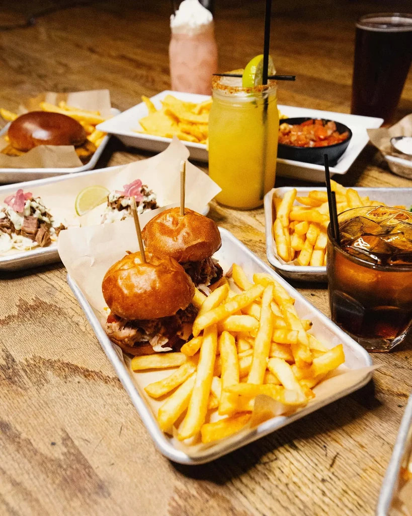 Overhead: Wooden table with burgers, fries, tacos, and multiple drinks.