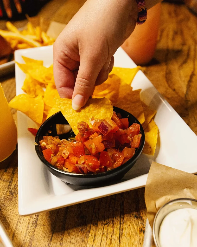 Hand dips yellow nacho chip into red salsa on a wooden table.