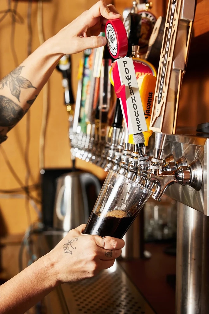 Tattooed hands pouring dark beer from a Powerhouse tap into a glass.