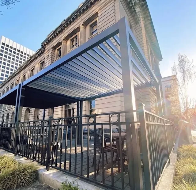 Empty urban patio with dark pergola, wicker furniture, classical building.