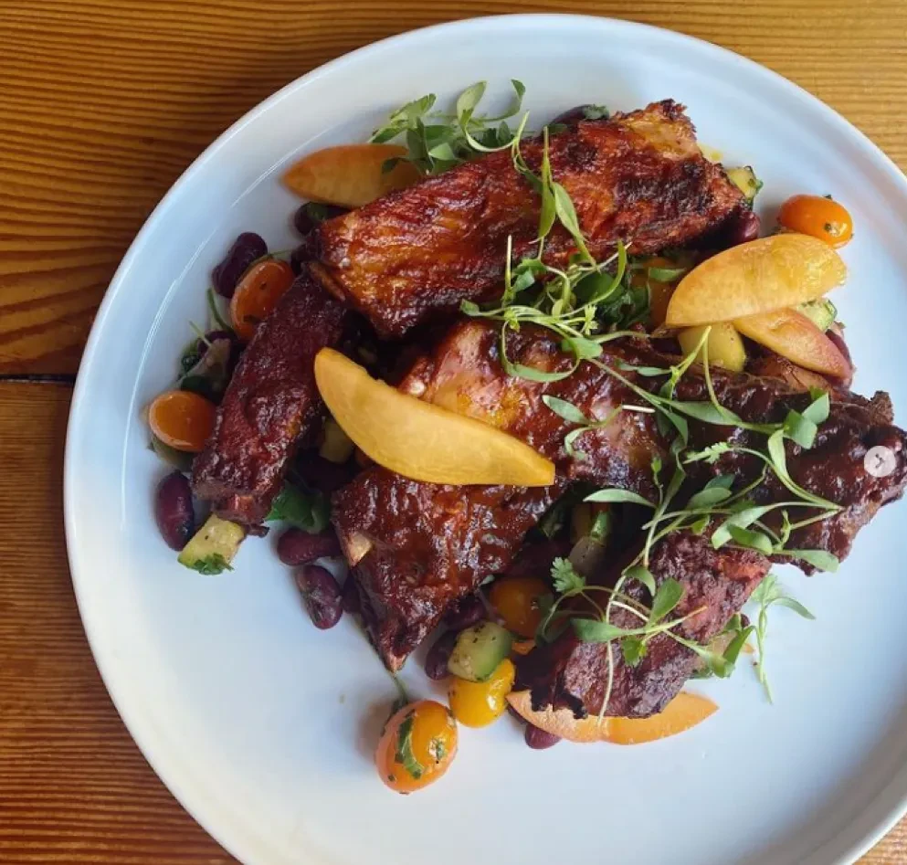 Glazed ribs, peaches, tomatoes, and greens plated on wood.
