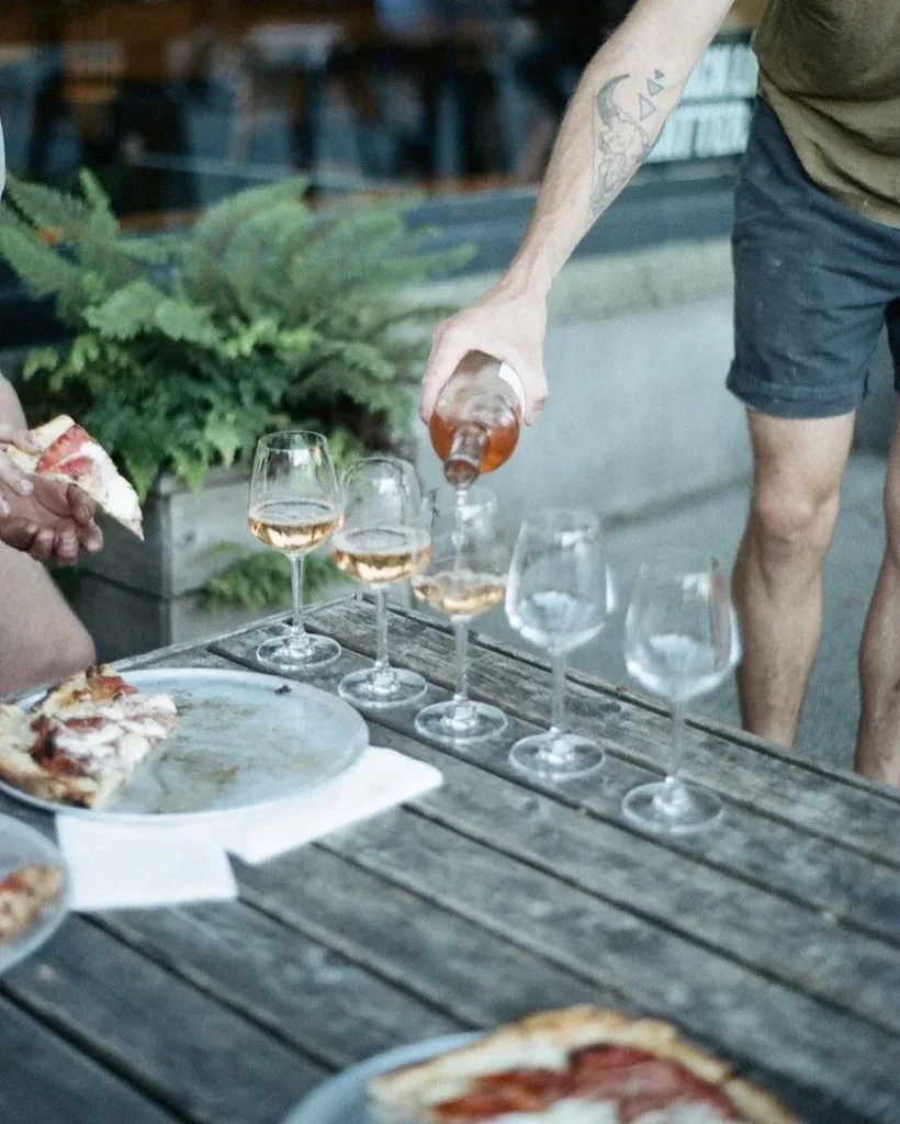 Person with tattooed arm pours rosé wine at an outdoor pizza meal.