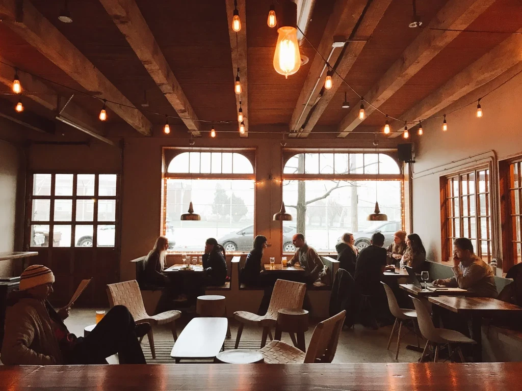 Restaurant interior with exposed beams, string lights, large arched windows, seated people.