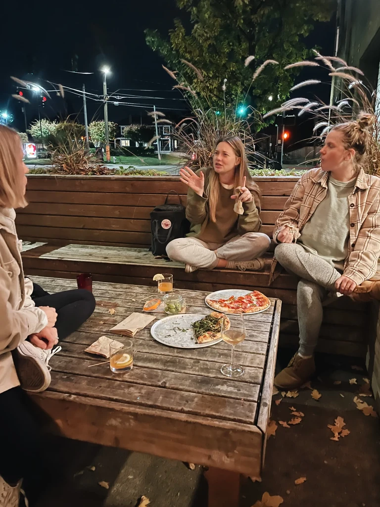 Three women at a nighttime outdoor table with pizza and drinks.