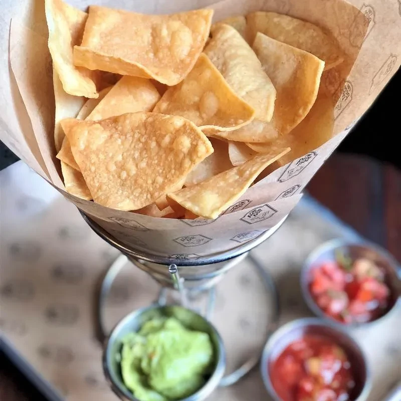 Golden tortilla chips in a branded cone with guacamole and two salsas.