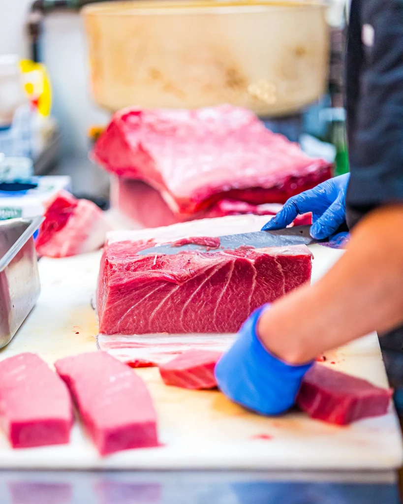Blue-gloved hand slicing raw red tuna on a white cutting board.