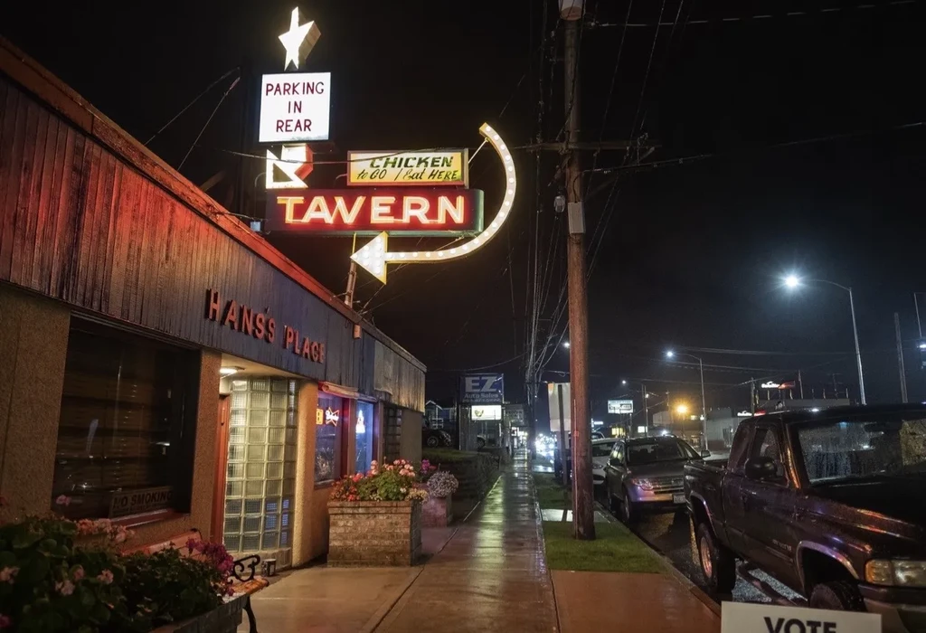 Hans's Place Tavern at night, neon signs reflecting on wet pavement.