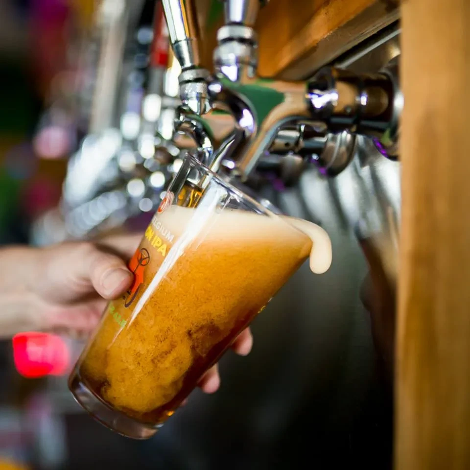 Amber beer pours from tap, overflowing a pint glass with foam.