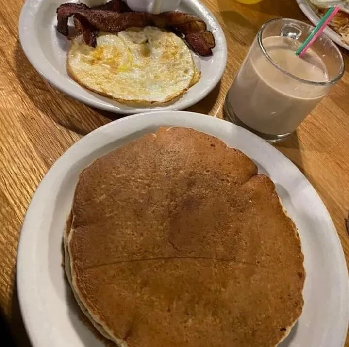 Breakfast of pancakes, fried eggs, bacon, and milk on wooden table.