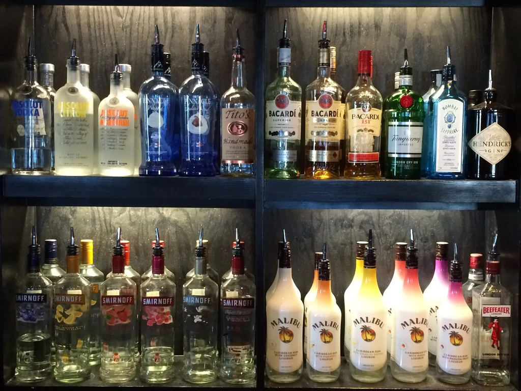 Assorted liquor bottles with pour spouts on dark bar shelves.