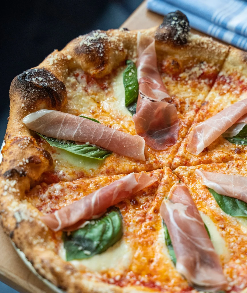 Wood-fired pizza, sliced, with prosciutto, mozzarella, and fresh basil.