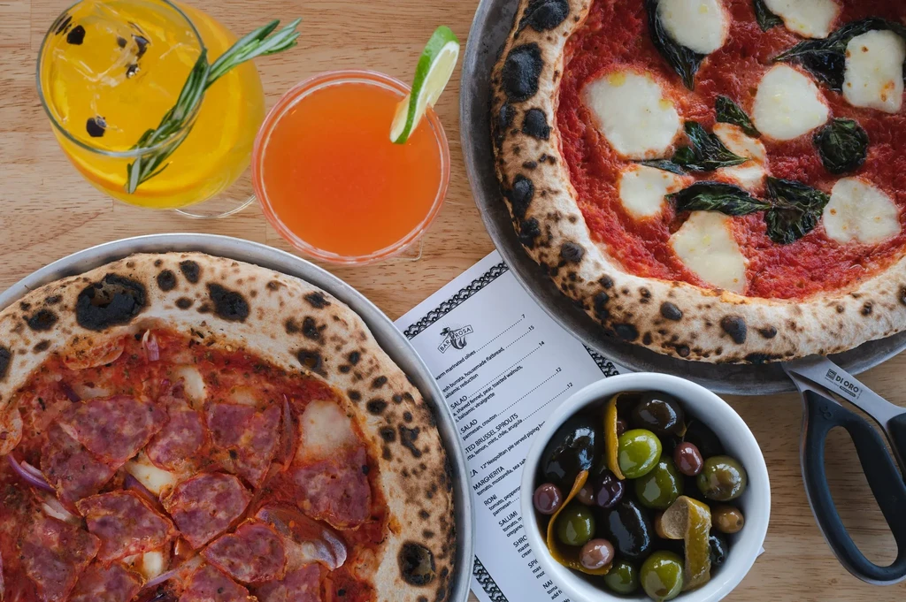 Overhead: two pizzas, cocktails, olives on a wooden table.