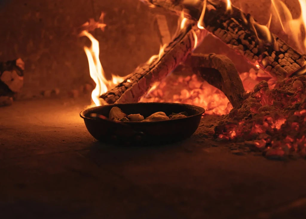 Food in cast-iron pan cooking in a fiery wood oven.