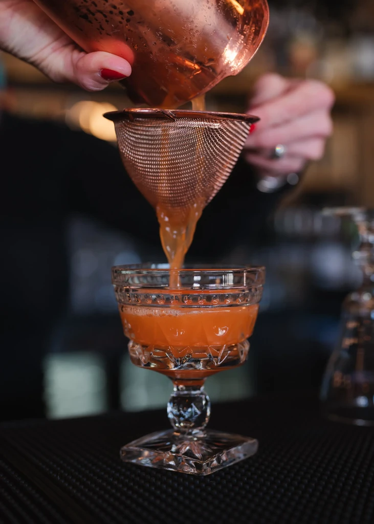 Amber cocktail strained from copper shaker into cut-glass goblet.