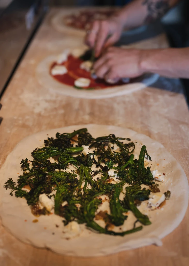Hands topping two pizzas with broccolini, cheese, sauce, and basil.