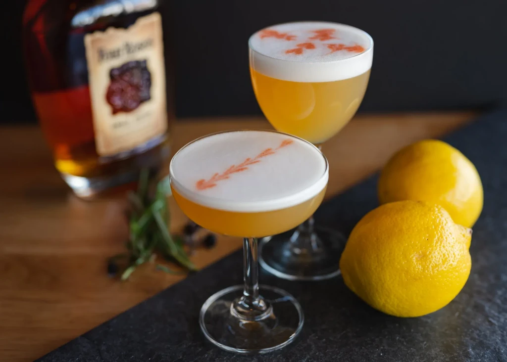 Two yellow cocktails with foam art, Four Roses bottle, lemons, berries.