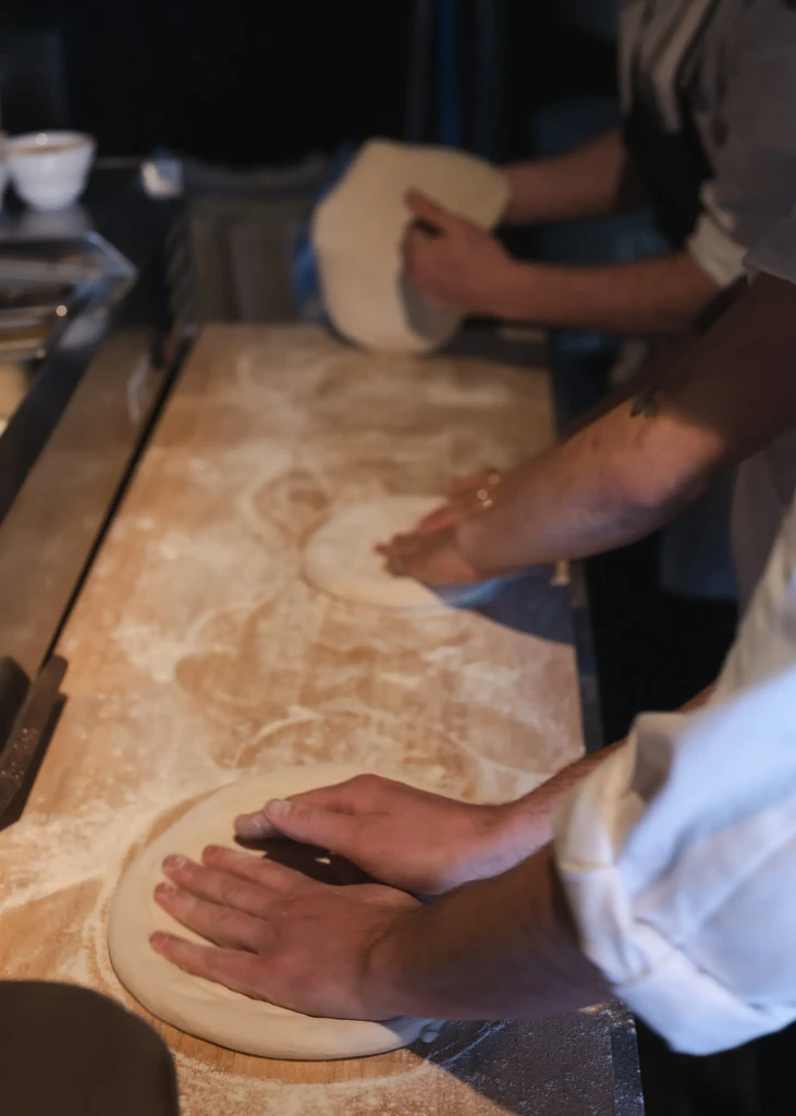 Multiple hands shape pizza dough on a flour-dusted wooden surface.
