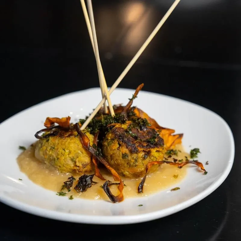Skewered golden-brown fritters with green interior, creamy sauce, herbs, and crisps.
