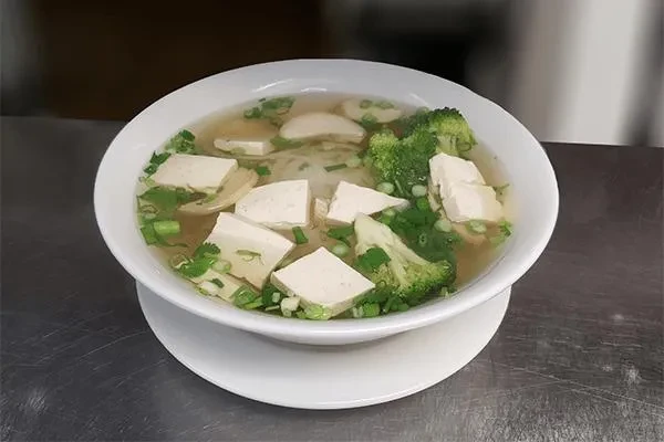 White bowl of clear broth soup with tofu, broccoli, green onions.