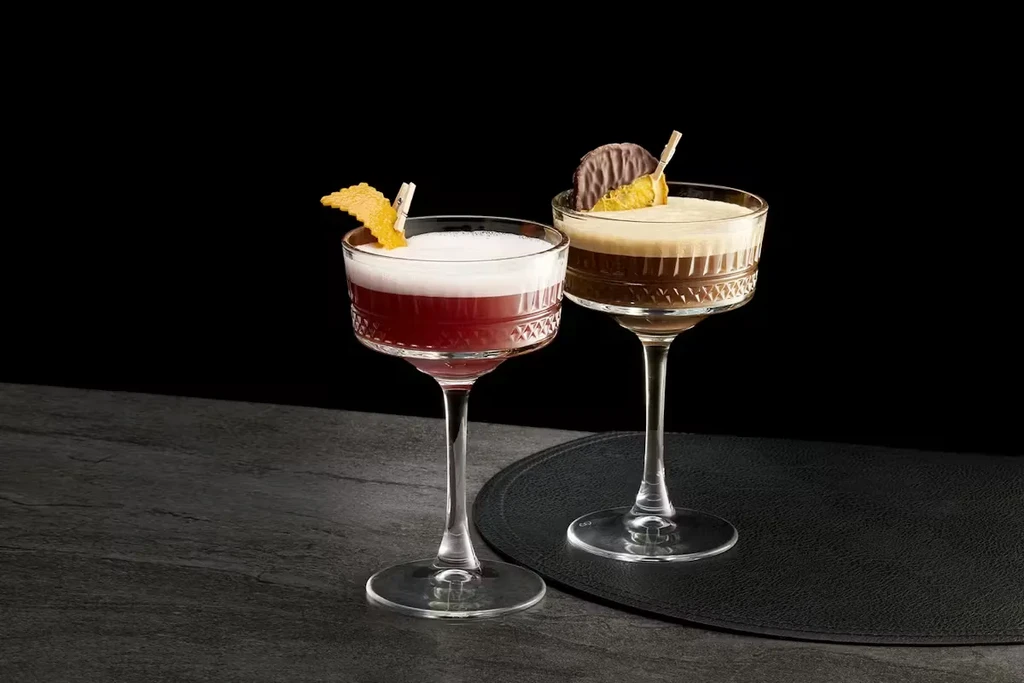 Two red and brown cocktails, uniquely garnished in cut-glass coupettes.