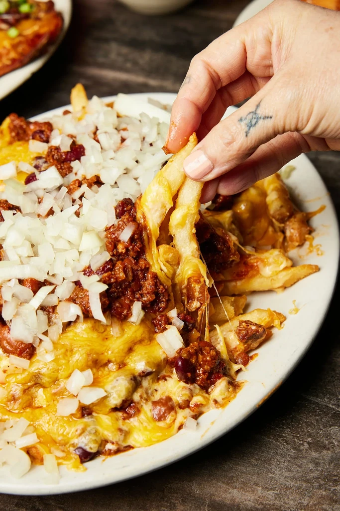 A hand pulls chili cheese fries with melted cheese and onions.