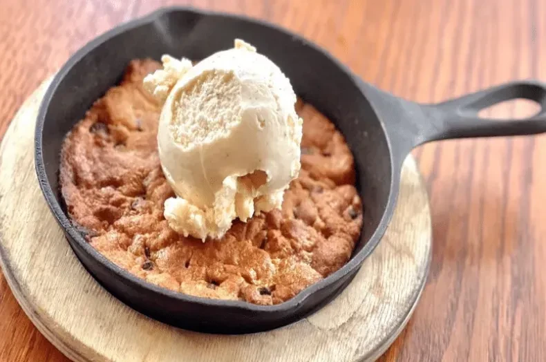 Warm chocolate chip cookie skillet, vanilla ice cream melting on wooden trivet.