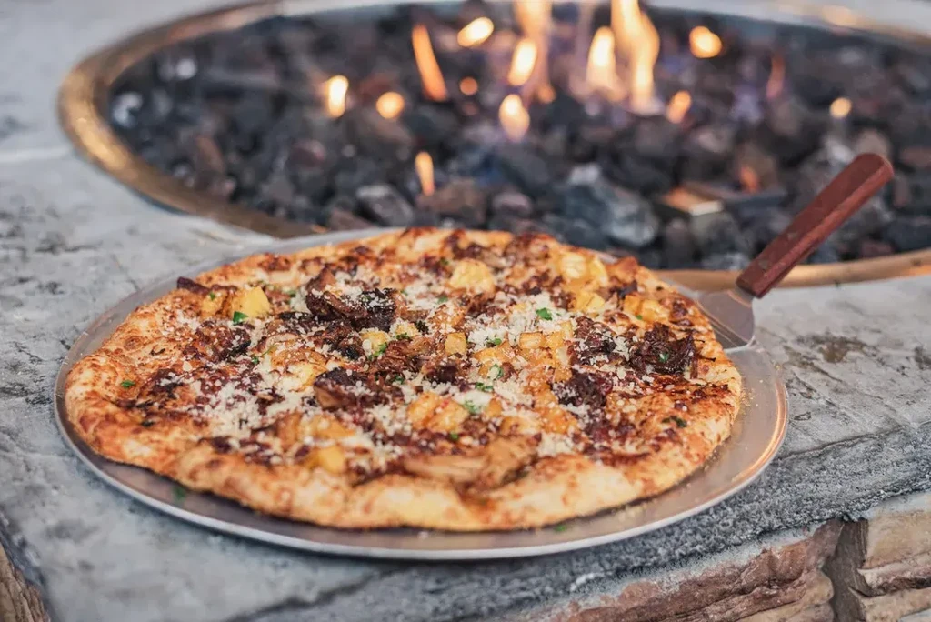 Meat, potato, cheese, and herb pizza on a pan by a fire.
